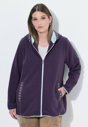 Žena stojící jednu ruku v kapse, oblečená do fialové fleecové bundy na zip s vzorovaným lemováním a kapucí, na bílém pozadí.