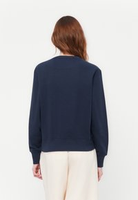 Frau mit langen braunen Haaren, die einen navyblauen Langarm-Pullover und hellbeige Hosen trägt, steht vor einem schlichten weißen Hintergrund.