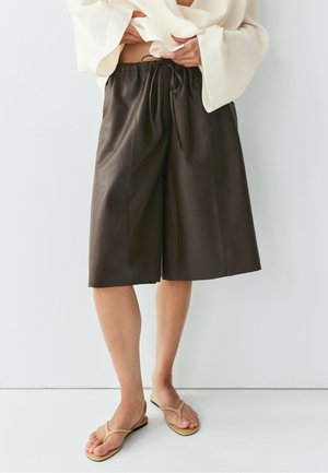 Person trägt schwarze Lederknielange Shorts, ein lockeres weißes Oberteil und beige Sandalen und steht vor einem hellen Hintergrund.