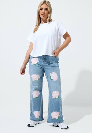 Frau trägt weißes T-Shirt und hellblaue Jeans mit großen pinken Rosenmotiven, steht mit einer Hand in der Hosentasche und weißen Sneakers.
