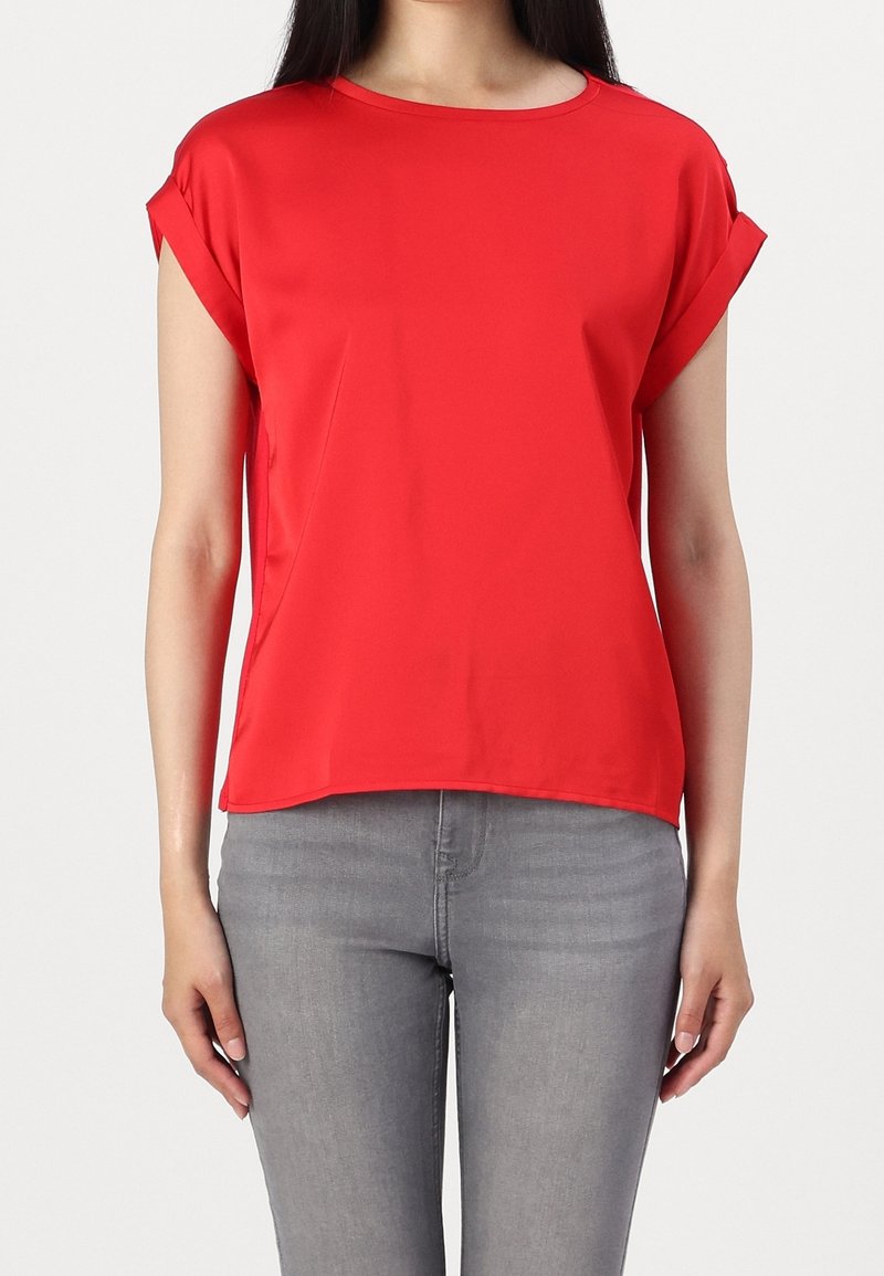 Femme portant un haut rouge vif à manches courtes avec des manches retroussées, assorti à un jean ajusté gris clair, sur fond uni.