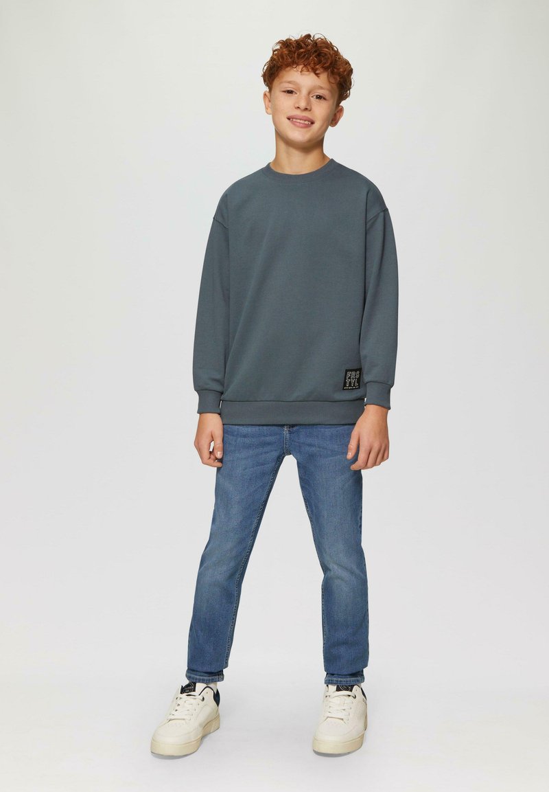 Grijze sweatshirt met geribde manchetten en zoom, label aan de linksonderzijde; gecombineerd met lichtblauwe jeans en witte sneakers.