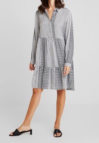 Robe rayée noir et blanc avec motif floral, boutonnée à l'avant, manches longues et jupe à volants. Associée à des chaussures noires sans lacets.