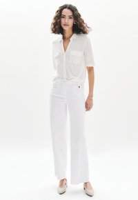 Camisa blanca de botones con mangas enrolladas y dos bolsillos en el pecho, combinada con pantalones de pierna ancha blancos de cintura alta y tacones de color claro.