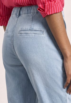 Osoba nosí světle modré džíny s vysokým pasem a zadními kapsami s lištou a červenou pruhovanou košili s vyhrnutými rukávy.