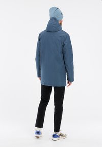 Veste imperméable bleue avec capuche, coupe décontractée et manches longues, accompagnée d'un pantalon noir et de chaussures de sport colorées.