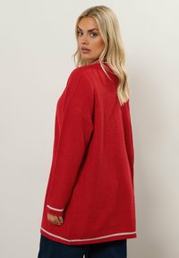 Röd stickad tröja med lös passform, försedd med vita randiga detaljer vid ärmslut och nederkant. Texturdesign med ribbad struktur och långa ärmar.