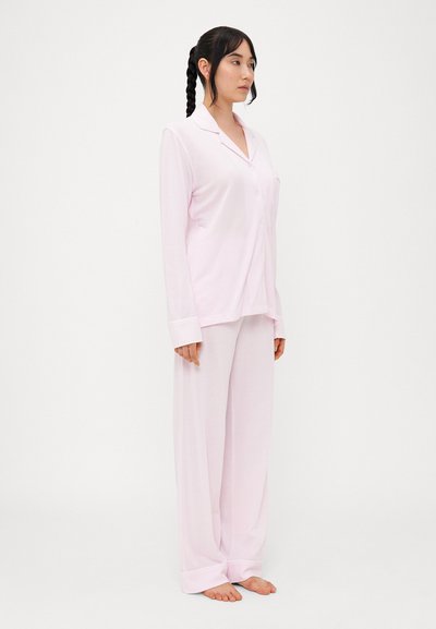 Ροζ πυτζάμα που περιλαμβάνει ένα φανελάκι με κουμπιά και μακριά μανίκια σε χαλαρή γραμμή και αντίστοιχες παντελόνες με φαρδιά πόδια, κατασκευασμένη από μαλακό, ελαφρύ ύφασμα.