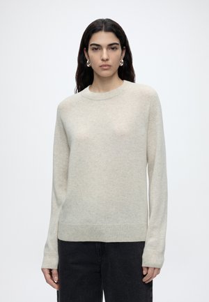 Donna con lunghi capelli scuri che indossa orecchini a cerchio color argento, un maglione beige chiaro con scollo rotondo e pantaloni neri, in piedi davanti a uno sfondo bianco.
