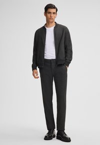 Giacca bomber nera con texture e polsini a coste, abbinata a pantaloni tapered coordinati e scarpe nere con lacci su uno sfondo neutro.