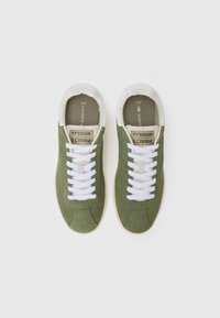 Sapatilhas em camurça verde com atacadores brancos, detalhes em creme e solas de borracha. Apresentam um rótulo na língua com a marca "TENNIS" e "LACOSTE".