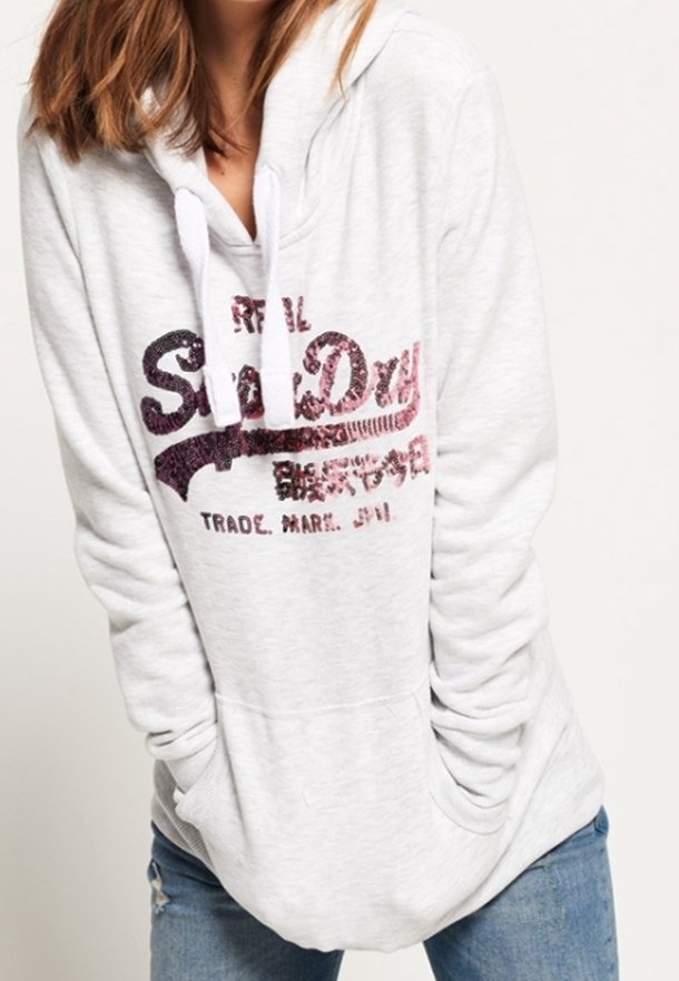 Femme portant un sweat à capuche blanc avec un texte pailleté rose et noir, mains dans les poches, associée à un jean bleu.