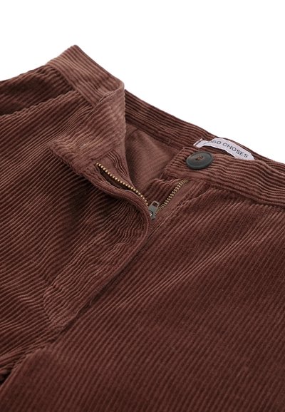 Gros plan d'un pantalon en velours côtelé marron vu de face, montrant une fermeture éclair partiellement ouverte, un bouton noir, une ceinture et une étiquette de marque visible à l'intérieur de la ceinture.