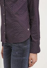 Chemise boutonnée violette foncée  à pois blancs, à manches longues et avec un ourlet arrondi. Présente un petit logo sur le côté inférieur.