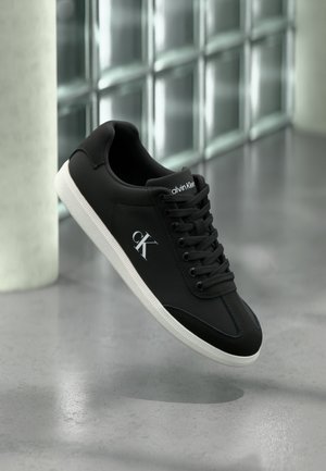 Czarne sneakersy o gładkiej teksturze, białej podeszwie i sznurowanej konstrukcji. Posiadają subtelne logo z boku oraz zaokrąglony czubek.