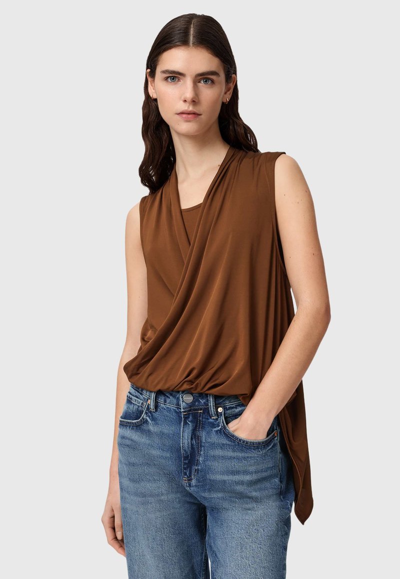 Vrouw met lang donker haar, gekleed in een mouwloze bruine top met drapering en blauwe spijkerbroek, met één hand in de zak, tegen een lichte achtergrond.