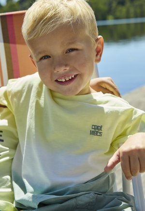 Light green and yellow gradient t-shirt with "Cool Vibes" text. Short sleeves, round neck, sitting by a lake. Shorts are muted green.