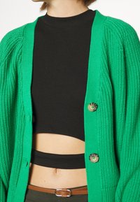 Zelený pletený kardigan s žebrovanou strukturou, s knoflíkovým zapínáním. Nosený přes černý přiléhavý crop top.