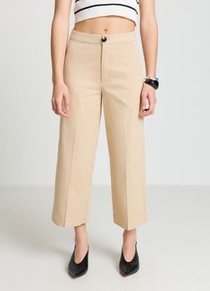 Pantalon beige à jambes larges avec fermeture à bouton, devant plat et sans poches. Associé à des talons noirs à bouts pointus, mettant en valeur une texture de tissu lisse.