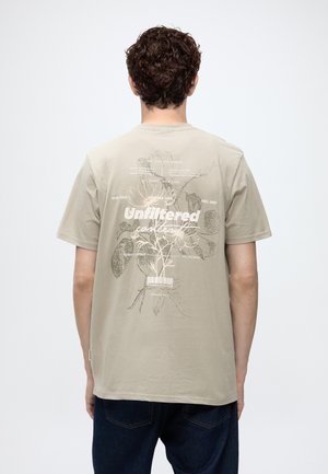 Mand med krøllet hår iført beige T-shirt med blomster- og tekstdesign, der lyder "Unfiltered collection" på bagsiden, stående mod hvid baggrund.