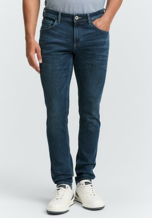Dark blue jeans with a slim fit, crafted from denim. Features pockets, a single front button, and metallic accents. Paired with white sneakers.