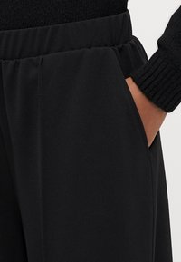 Pantalons noirs en tissu avec une texture douce, comportant une taille élastique et des poches latérales, partiellement visibles contre un pull noir.