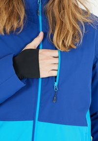 Persona con i capelli lunghi che indossa una giacca blu mentre apre la tasca del petto, rivelando un guanto nero con un foro per il pollice sotto.