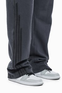 Grå sweatpants med tre sorte striber langs siden, der har en løs pasform og blød tekstur, kombineret med hvide og grå sneakers.