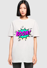 Balta didelis t-shirt'as su spalvinga komiksų stiliaus grafika, kurioje žodis "BOOM!" pavaizduotas rožine ir mėtų spalvomis, supaprastintas juodais kontūrais ir taškeliais.