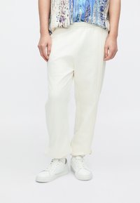 Białe bawełniane spodnie dresowe o luźnym kroju, z plisami po bokach, elastycznym paskiem w talii, zestawione z białymi trampkami.