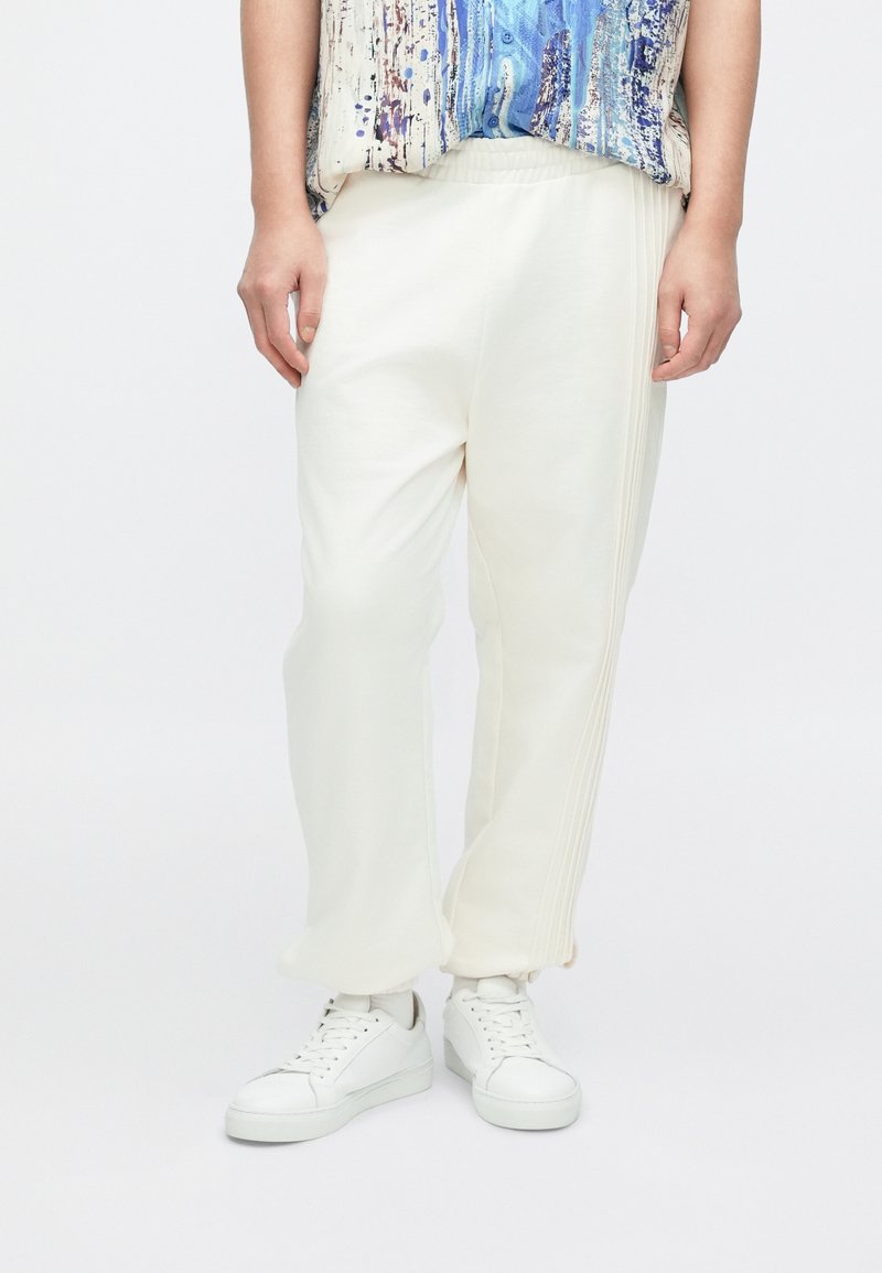 Białe bawełniane spodnie dresowe o luźnym kroju, z plisami po bokach, elastycznym paskiem w talii, zestawione z białymi trampkami.