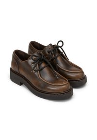 Une paire de chaussures en cuir marron à lacets avec de épaisses semelles noires, présentant une couture moc toe et des œillets en métal sur un fond blanc.