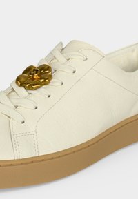 Baskets en cuir blanc avec une semelle beige, des lacets blancs et un accessoire de lacet doré en forme de fleur avec un emblème circulaire.
