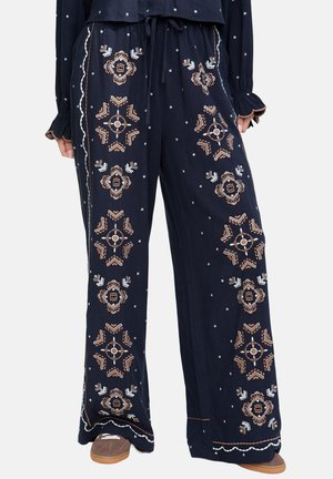 Marineblaue Palazzohose mit beigem und weißem geometrischem Stickmuster, Kordelzugbund, getragen mit passendem Oberteil und braunen Sneakern.