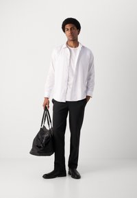 Un sac de sport noir tenu d'une main, porté avec une chemise blanche superposée sur un t-shirt blanc, associé à un pantalon sombre et des chaussures noires.