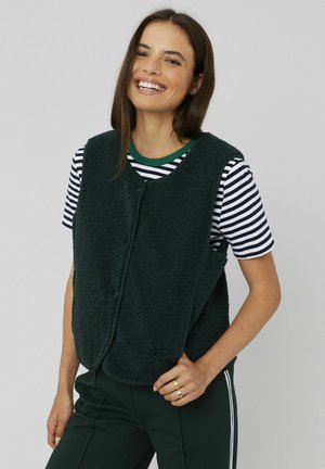 Femme souriante portant un gilet en polaire noir sur un t-shirt striped noir et blanc, un pantalon sombre avec des bandes latérales blanches et des bagues en or sur sa main droite.