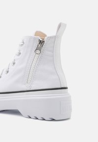 Białe trampki na wysokiej cholewce z zamkiem bocznym, czarny akcent na podeszwie oraz teksturowana gumowa podeszwa dla lepszej przyczepności. Widoczne logo Converse.