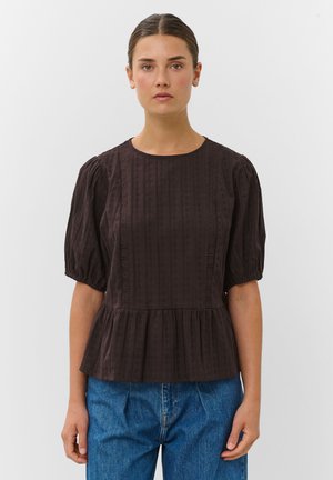 Jeune femme portant un chemisier texturé marron foncé avec des manches bouffantes et un jean bleu sur un fond blanc uni.
