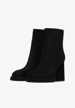 Botine negre din suede, cu o textură netedă, vârf rotund, toc înalt cub și închidere cu fermoar pe lateral. Talpa prezintă un contrast ușor.