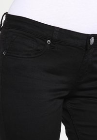 Jeans de mezclilla negra con una textura suave, que cuentan con un botón plateado y bolsillos frontales. Vista cercana de la cinturilla y los detalles de la tela.