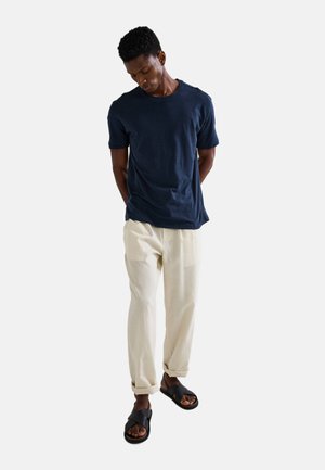 T-shirt bleu marine à manches courtes, pantalons crème plissés et sandales noires. La tenue présente une coupe décontractée et des textures de tissu légères.