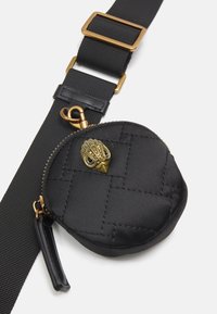Kulatá černá prošívaná taštička vyrobená z saténu se zipovým zapínáním, s dekorativním zlatým přívěskem ve tvaru ruky a připevněná na širokém černém popruhu.