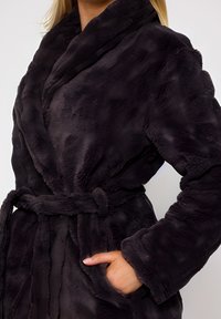 Kvinde iført sort faux fur frakke med bælte, hånd i lommen, vist fra hals til hofte på en hvid baggrund.