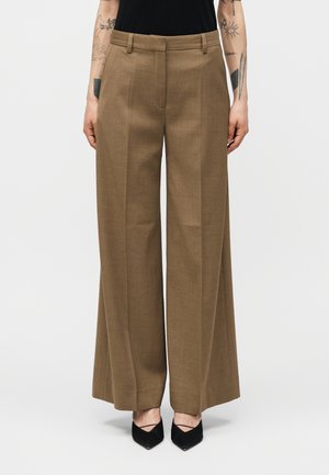 Γυναίκα που φοράει καφέ παντελόνια με φαρδύ πόδι και πιέτες μπροστά, καθώς και μαύρες μυτερές γόβες, στέκεται μπροστά σε ένα απλό φόντο.