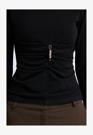 Schwarzes Langarmoberteil mit gerafftem Stoff und einem goldenen Sicherheitsnadeldetail, das in der Mitte mit klaren Edelsteinen verziert ist. Glatte Oberfläche, figurbetontes Design.