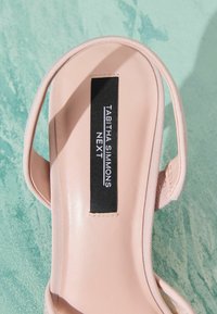 Gros plan sur une semelle intérieure de sandale rose pâle avec une étiquette noire indiquant « Tabitha Simmons NEXT » sur une surface turquoise texturée.