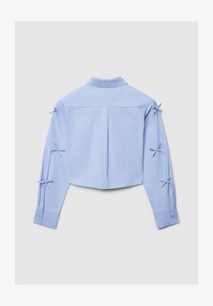 Camicia a maniche lunghe corta azzurro chiaro con colletto e fiocchi decorativi annodati su entrambe le maniche, vista da dietro.
