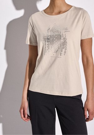 Femme portant un t-shirt beige à manches courtes avec un motif abstrait de skyline et un pantalon noir sur un fond clair uni.