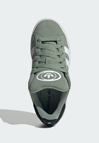 Zelené semišové tenisky s bielymi pruhmi, okrúhlym logom na jazyku a textúrovaným interiérom. Obsahujú čierny akcent na päte.