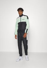 Träningsoverall i mintgrön och svart, med en zip-up jacka med kontrasterande paneler och matchande smalna byxor. Buren med vita sneakers.